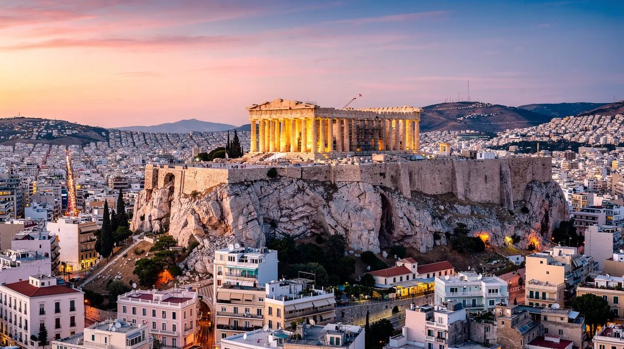 Imagem de Atenas, mostrando a famosa Acrópole ao fundo, com suas antigas colunas e ruínas, cercadas por montanhas e a cidade moderna ao redor. Esta imagem representa um destino popular para viagens, com diversas opções de voos e pacotes promocionais disponíveis para turistas do Brasil e do mundo.