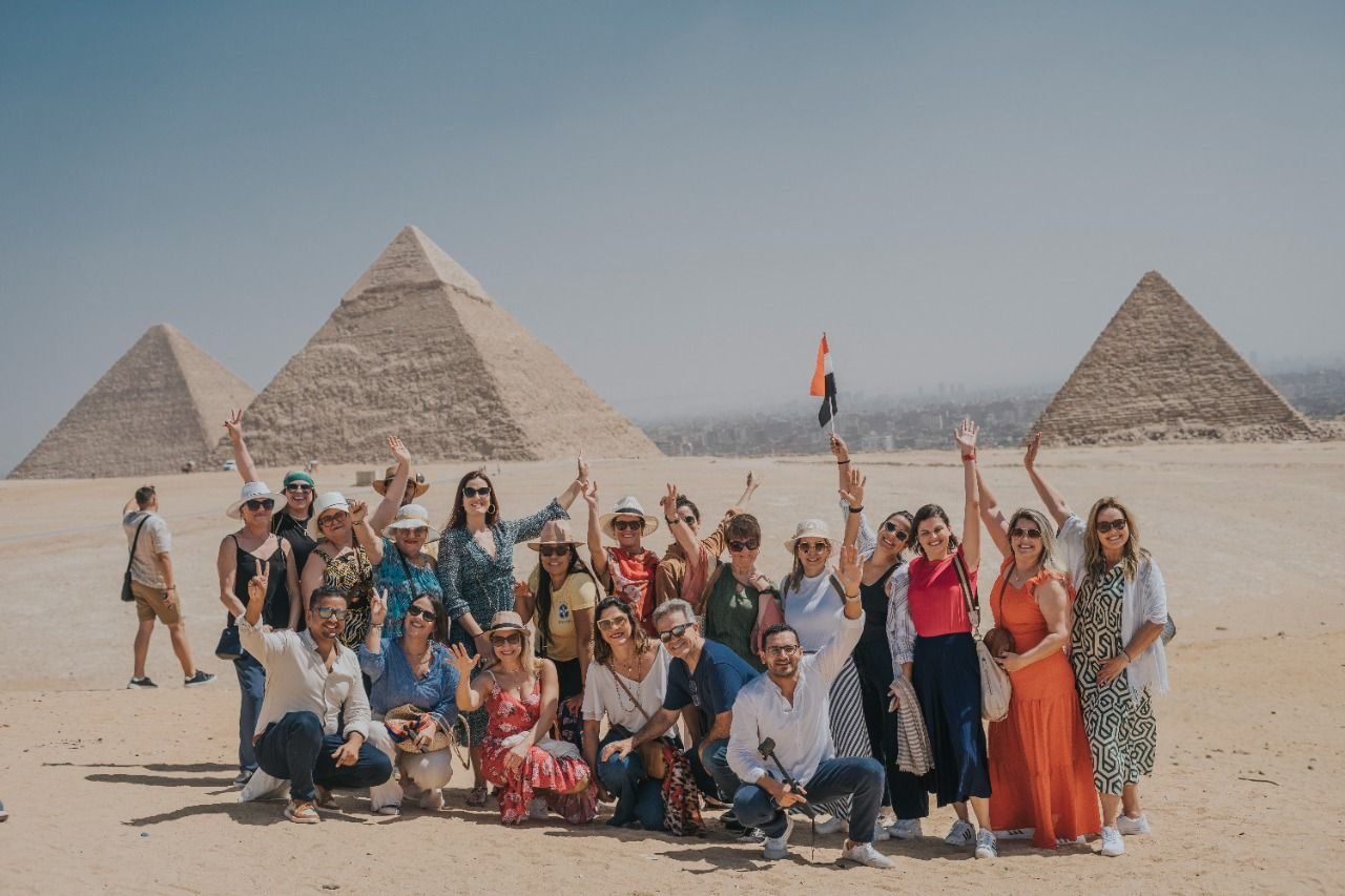 Grupo de clientes da EGP Viagens comemorando com as mãos para o alto e bandeira do Egito em frente às três Pirâmides de Gizé