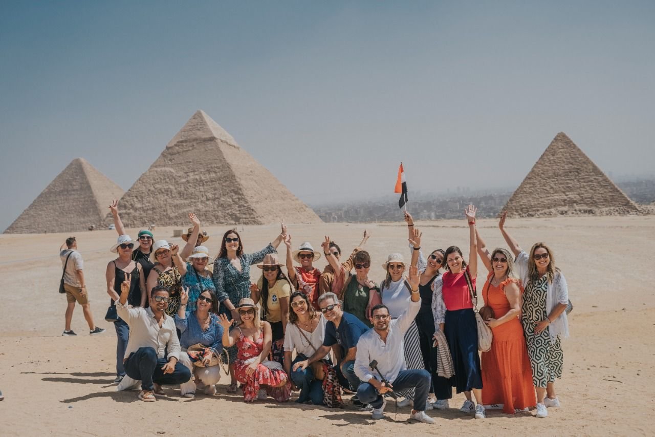 Grupo de clientes da EGP Viagens comemorando com as mãos para o alto e bandeira do Egito em frente às três Pirâmides de Gizé